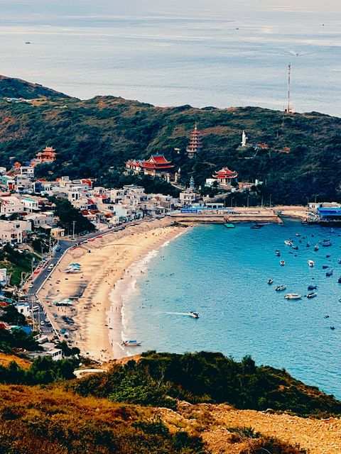 Curved sandy bay below a green hill with red-roofed pagodas, boats on turquoise water — shot on ELEV MAPLE