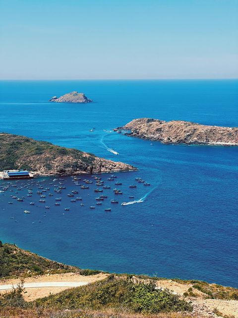 Blue bay dotted with fishing boats between rocky islets, seen from a grassy hillside — shot on A NFX